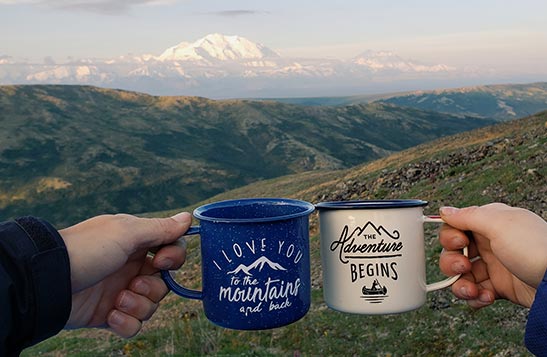 Two enamel coffee cups in a cheers in front of a wide tundra vista.