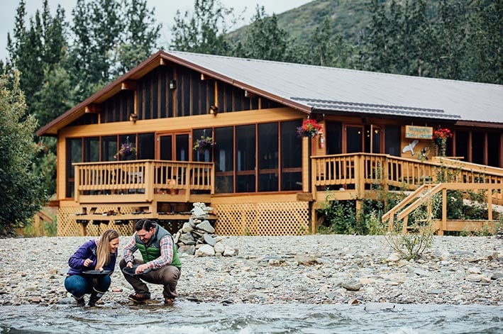 Two people pan for gold next to a wooden lodge.