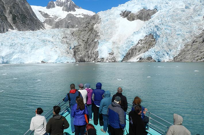 Boat guests stand at the prow of a boat before a tidewater glacier.