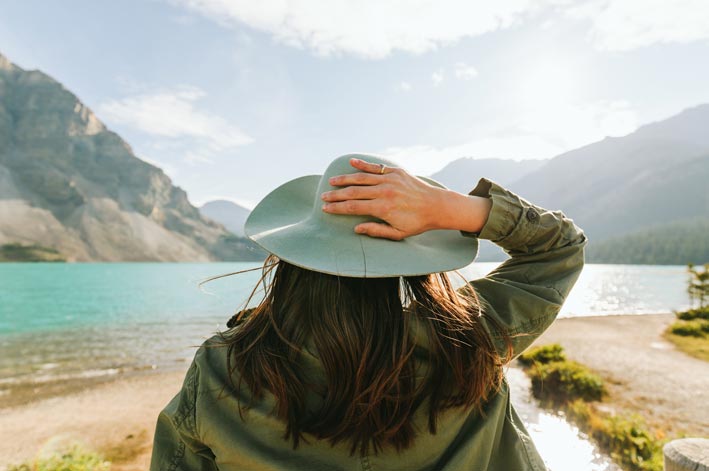 A woman holds onto her hat while looking towards a blue mountain lake