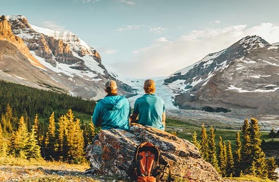 Two people sit on a rock looking towards mountains and glaciers.