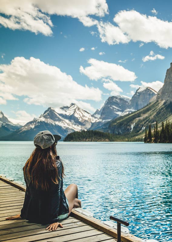 A person sits on a dock edge looking out across a blue lake towards mountains