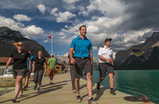A group of boat and deck crew walk on a deck along a turquoise lake.