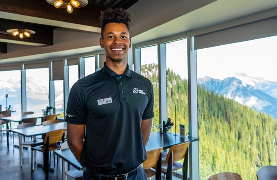 A worker smiles in a dining room that overlooks a forested mountainside.