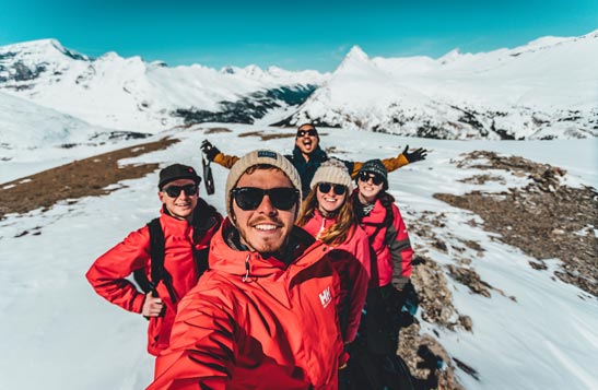Five hikers smile for a wide-angle selfie in a rocky and snowy alpine environment.