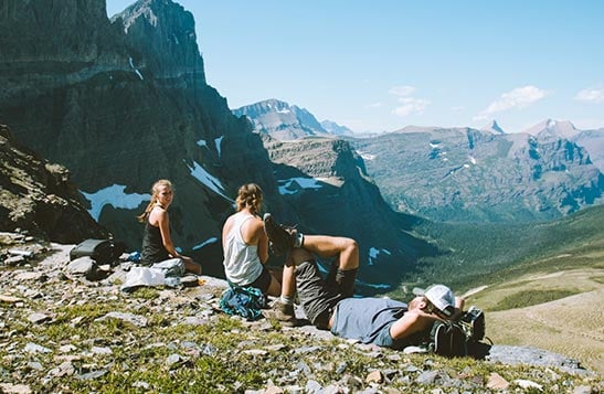 Three people sit in a rocky alpine meadow above a wide valley.