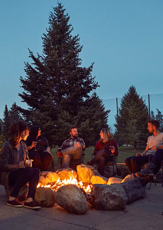 A group of six people sit around a firepit on a dusky evening.
