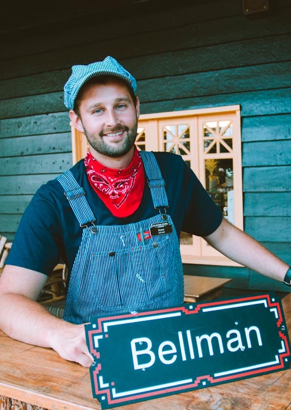 A bellman dressed in vintage-style overalls and hat smiles at the outdoor bellman desk.