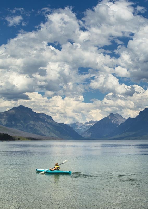 A kayaker paddles on a large lake surrounded by tree-covered mountains
