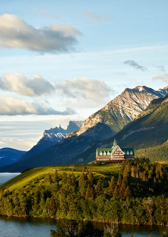 The Prince of Wales Hotel sits on a hill above a lake surrounded by mountains.