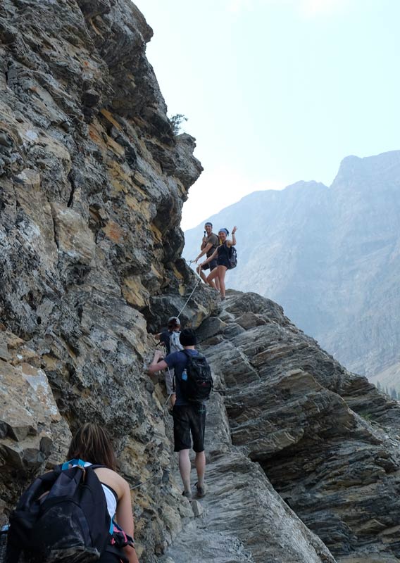 A group of hikers walk along a narrow path, holding onto a safety cable.