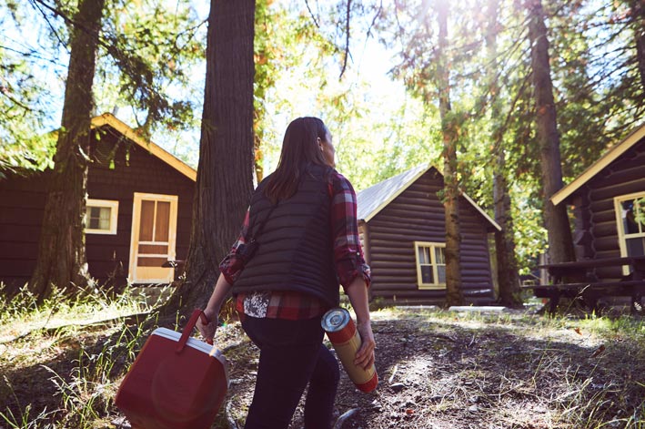 A woman walks under tall conifers between wooden cabins.