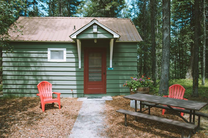 A green wooden cabin surrounded by trees with red chairs out front