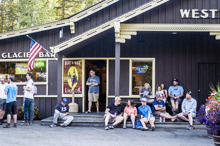 A crowd of people sit on and around a covered porch out front of the West Glacier Bar