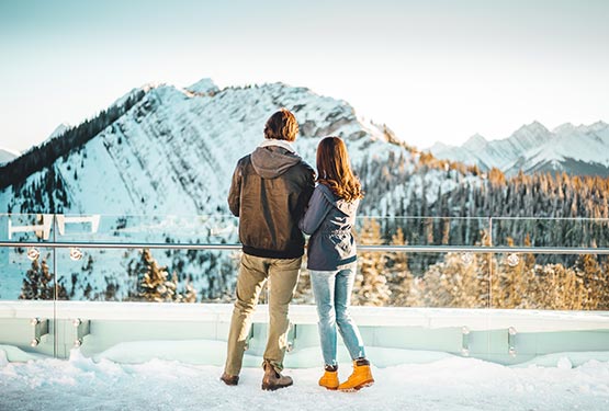Two people stand at a viewpoint looking towards a snow-covered mountain ridge.